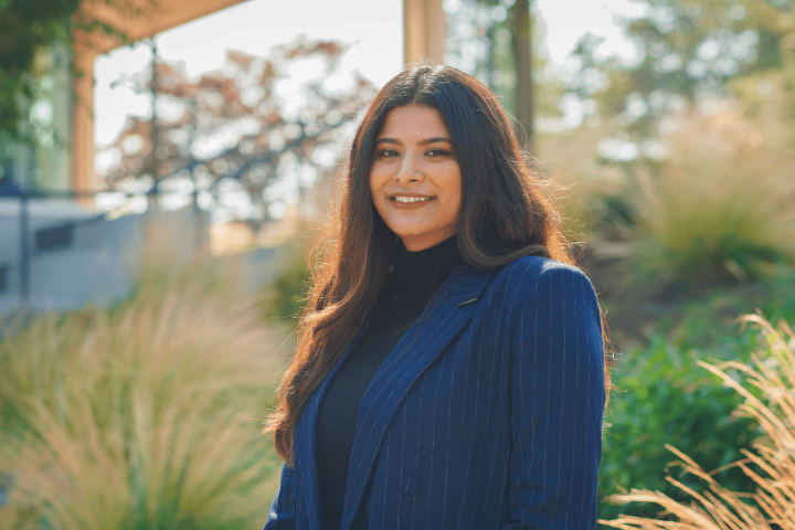 Professional woman with long, dark hair wearing a navy blue pinstripe blazer and black turtleneck, smiling outdoors in a garden setting with soft sunlight and blurred greenery in the background.